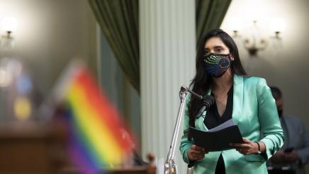 Assemblymember Cervantes wearing a green coat and LGBTQ Pride Mask Speaks in Support of HR 51 Declaring June as LGBTQ Pride Month
